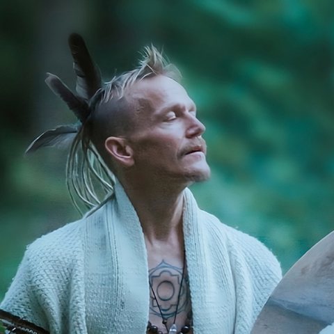 Mikael Ananda Cromsjö, spiritual teacher, with feathers in his hair, eyes closed, playing a shaman drum in the forest